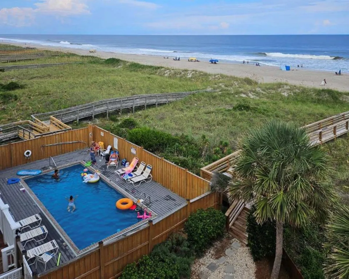 A vibrant oceanfront community pool surrounded by lush greenery, poolside loungers, and a clear view of the beach and boardwalk nearby. Families and children can be imagined swimming and playing in this joyful, active setting.