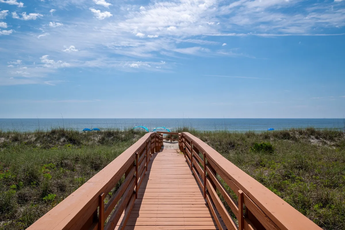 A long wooden boardwalk stretches across dune grass towards the vast, open ocean. The scene is serene and offers a direct, private pathway from the property to the pristine beach, with the promise of ocean breezes and easy beach access.
