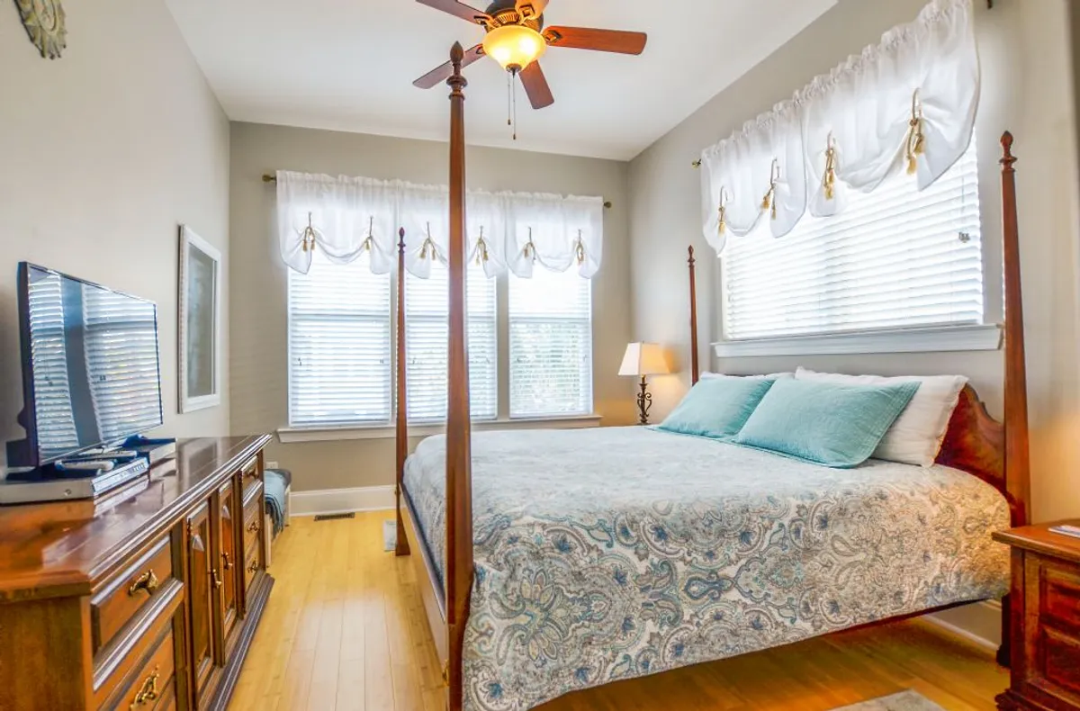 A tranquil bedroom featuring a four-poster king bed adorned with decorative pillows, large sun-filled windows, a wooden dresser, and a ceiling fan. The soft natural light and serene decor evoke a sense of peaceful retreat, making it ideal for restful escapes.