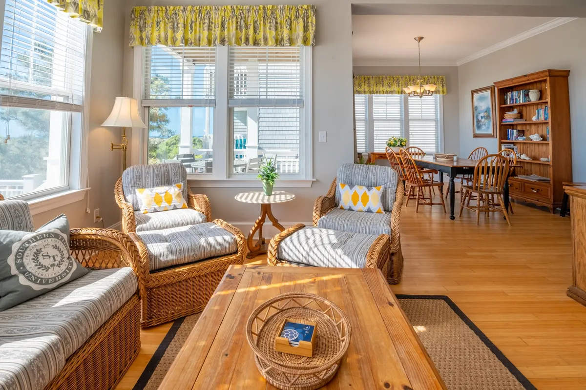 A warm, inviting living and dining area with wicker chairs, a wooden coffee table, and a dining table in the background—all set beside large windows that let in natural light and showcase the ocean beyond. The layout encourages both relaxation and social activities like meals and board games.