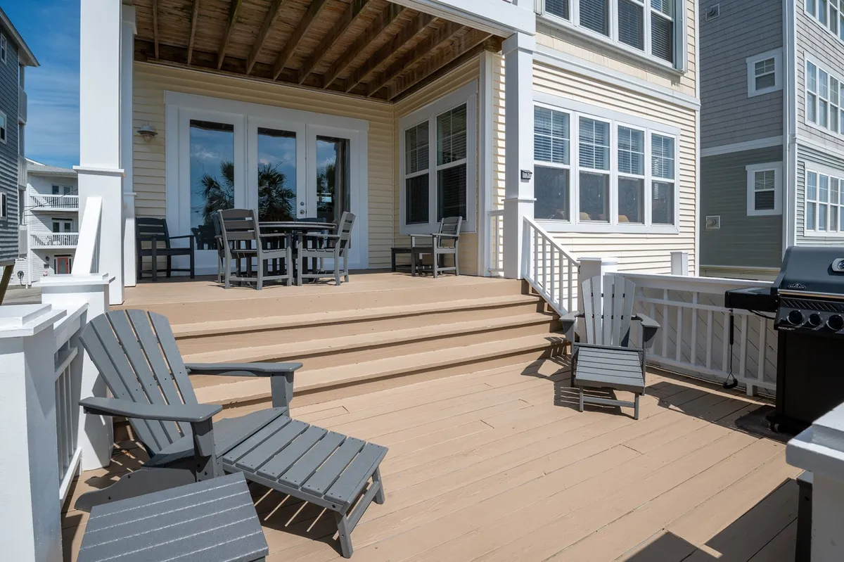An inviting outdoor patio featuring a spacious dining table, Adirondack chairs, a grill, and steps leading to a lush yard. The setting is ideal for alfresco meals and gatherings, surrounded by greenery and open sky.