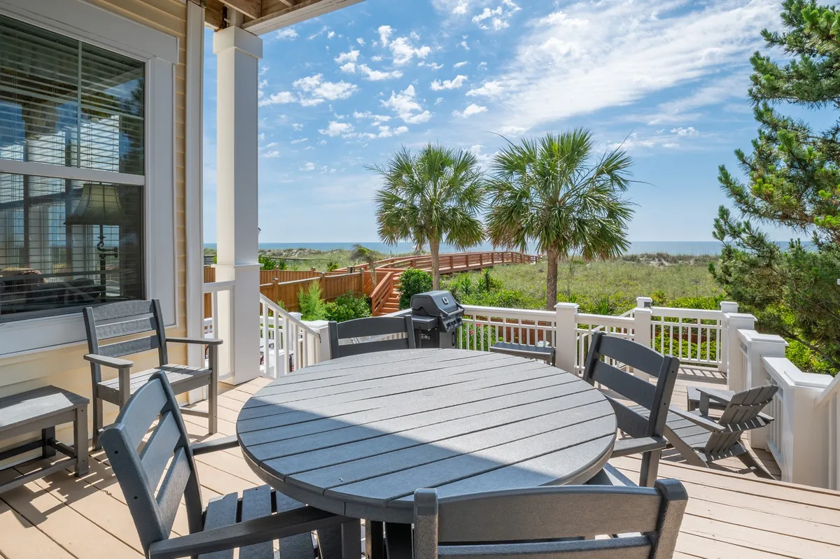 An oceanfront deck furnished with an outdoor dining table, surrounded by palm trees and overlooking a beautiful ocean view. A wooden walkway leads toward the beach, and the setting invites grilling, dining, and relaxing with family and friends in a cheerful, coastal atmosphere.