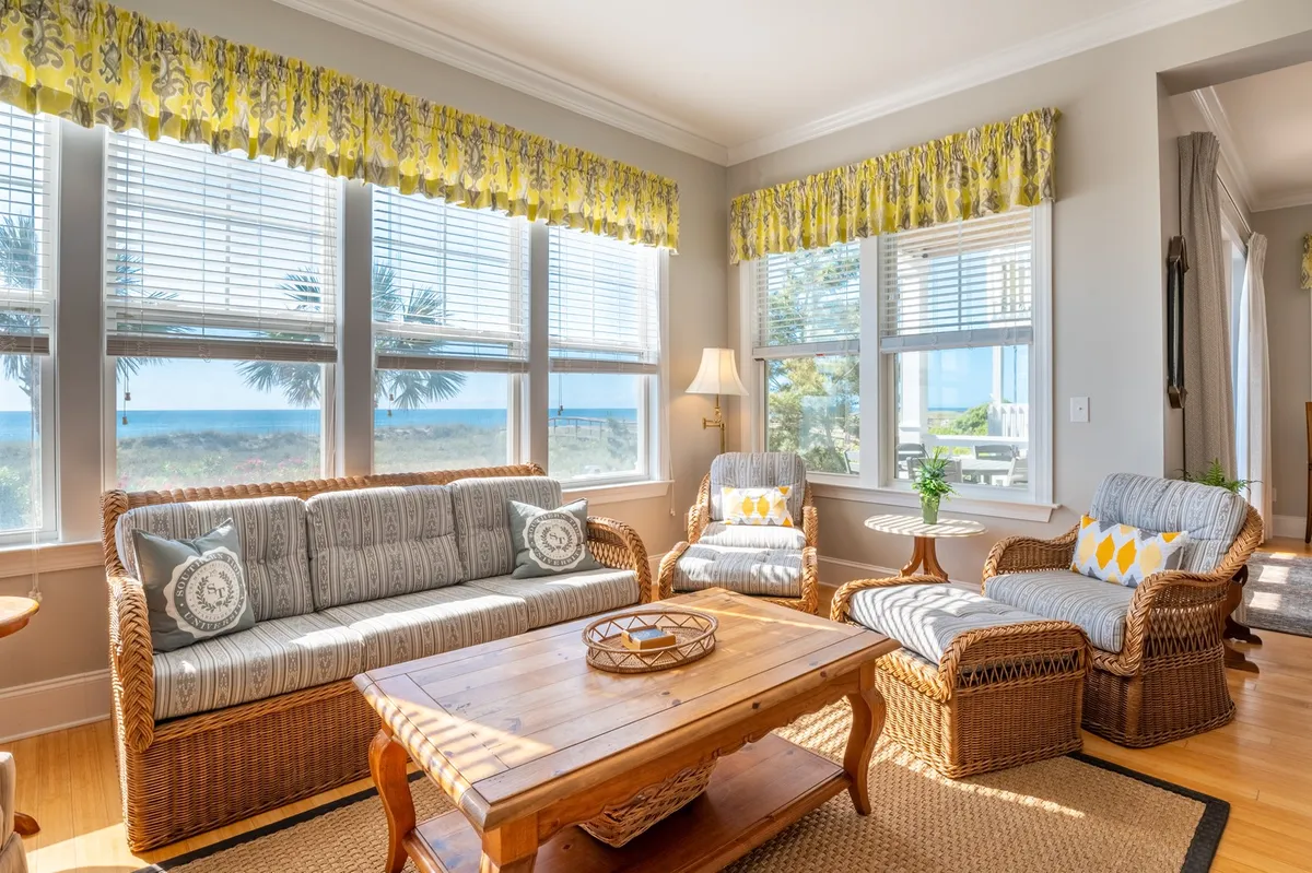 A cozy living room with a large sofa, coffee table, and expansive windows that frame sweeping beach views. The sunlight pouring in creates a serene, inviting environment perfect for relaxing after engaging in outdoor adventures.