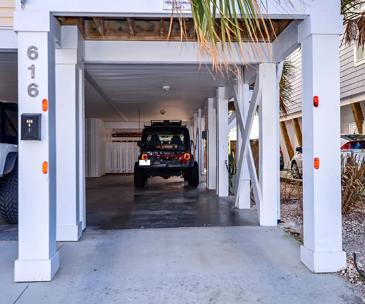 The image shows a tandem parking area beneath the duplex, featuring a black vehicle parked under white structural beams, with a numbered mailbox and decorative palm leaves in the background. The sheltered space is practical for families and groups arriving for their stay, and the marked parking spots provide clear organization.