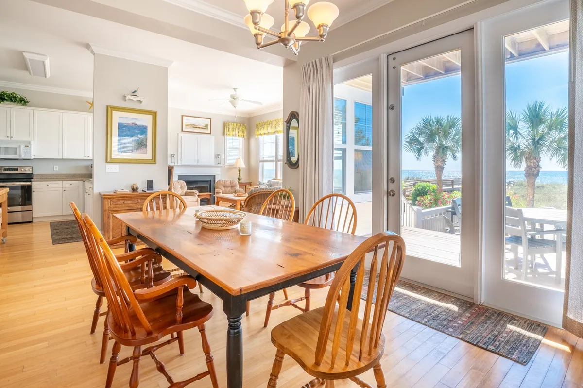 A bright, welcoming dining area centered around a wooden table, surrounded by large windows that showcase swaying palm trees and glimpses of the ocean just beyond. The kitchen is visible nearby, inviting gatherings for meals, laughter, and togetherness—all under the warm Carolina Beach sun.