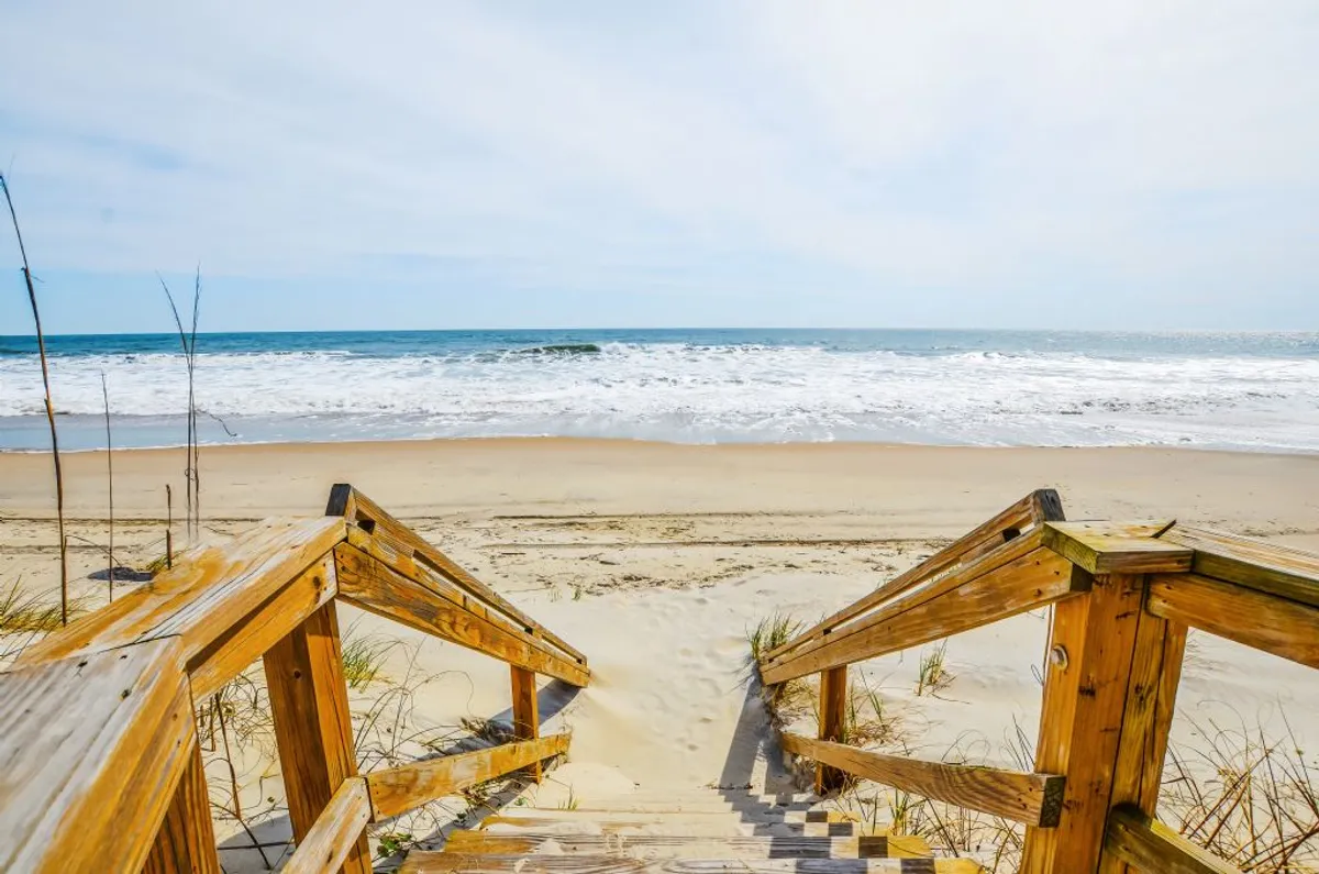 Wooden stairs lead directly onto the sandy beach, with gentle ocean waves visible in the background. The atmosphere is peaceful and inviting, suggesting quiet strolls along the shore, sunbathing, or simply watching the water.