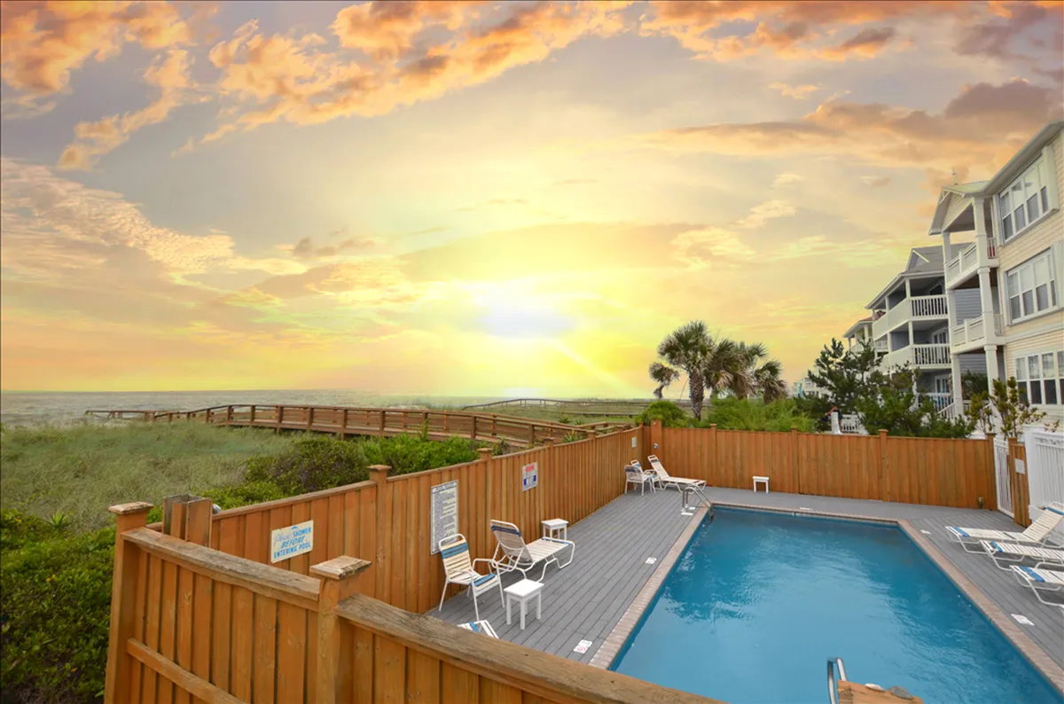 This exterior scene captures a stunning community swimming pool with a wooden fence, lounge chairs, and a pathway that leads directly toward the beach. The pool is bathed in the warm colors of sunset, and the tranquil water is inviting for swimmers and sunbathers alike. Palm trees add to the relaxed, resort-style atmosphere.