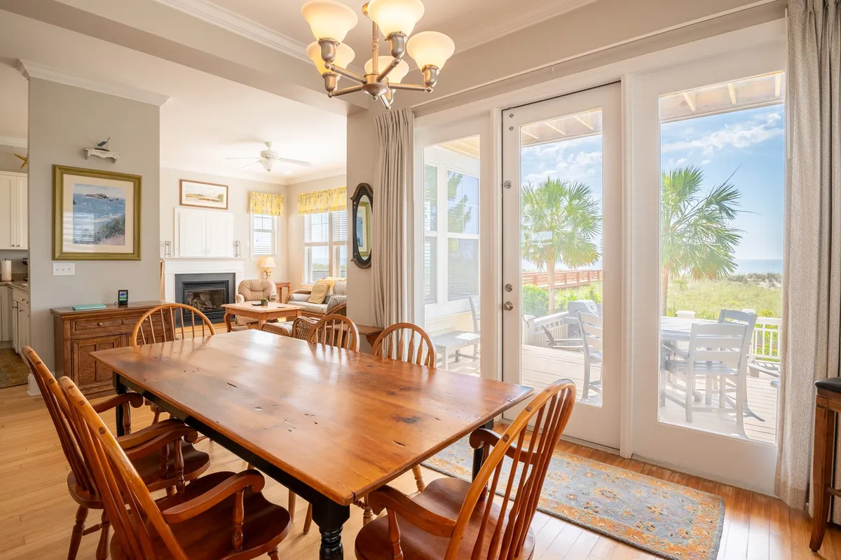 An elegant dining area with a wooden table, sliding glass doors that open to palm trees and the ocean, and a fireplace in the background. The space is filled with natural light, offering a seamless transition between indoor dining and outdoor enjoyment.